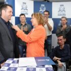 Mulher de casaco laranja sorri enquanto coloca pin no blazer de homem, outras pessoas aplaudem ao redor