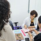 Mulher de jaleco branco está sentada à frente de mulher vestida de preto, as duas manuseiam um caderno, enquanto uma menina em pé assiste à cena