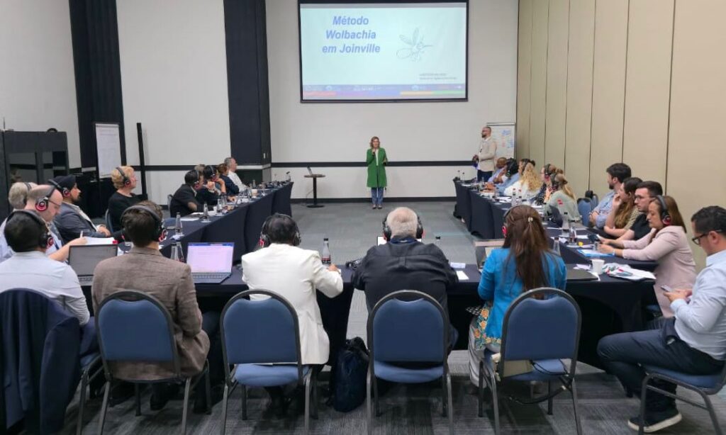 Mulher loira vestida de verde está em pé com microfone à frente de telão que diz Método Wolbachia em Joinville enquanto pessoas sentadas em mesa em forma de U assistem à apresentação
