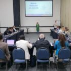 Mulher loira vestida de verde está em pé com microfone à frente de telão que diz Método Wolbachia em Joinville enquanto pessoas sentadas em mesa em forma de U assistem à apresentação