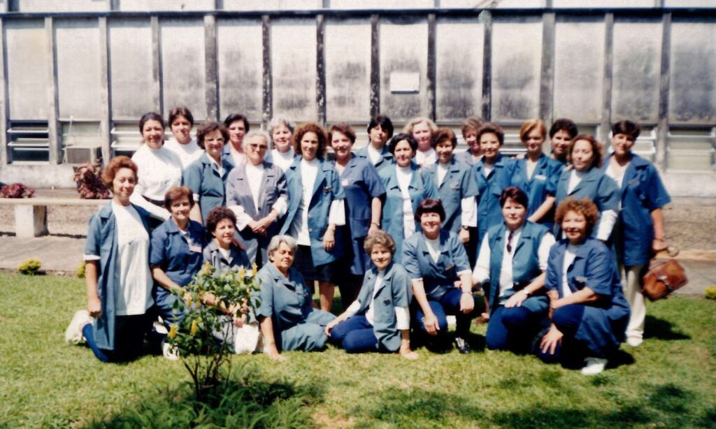 Grupo de voluntárias com mulheres vestidas com jaleco azul posa para foto em gramado perto de prédio