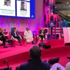 Quatro homens e uma mulher estão sentados em cadeiras em um palco, a mulher está com o microfone