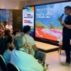 Homem com microfone está em pé, perto de telão, enquanto pessoas sentadas assistem à apresentação