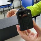 Imagem mostra uma mão segurando uma câmera corporal, um dispositivo preto