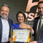 Mulher entre dois homens segura quadro enquanto todos olham sorrindo para a câmera