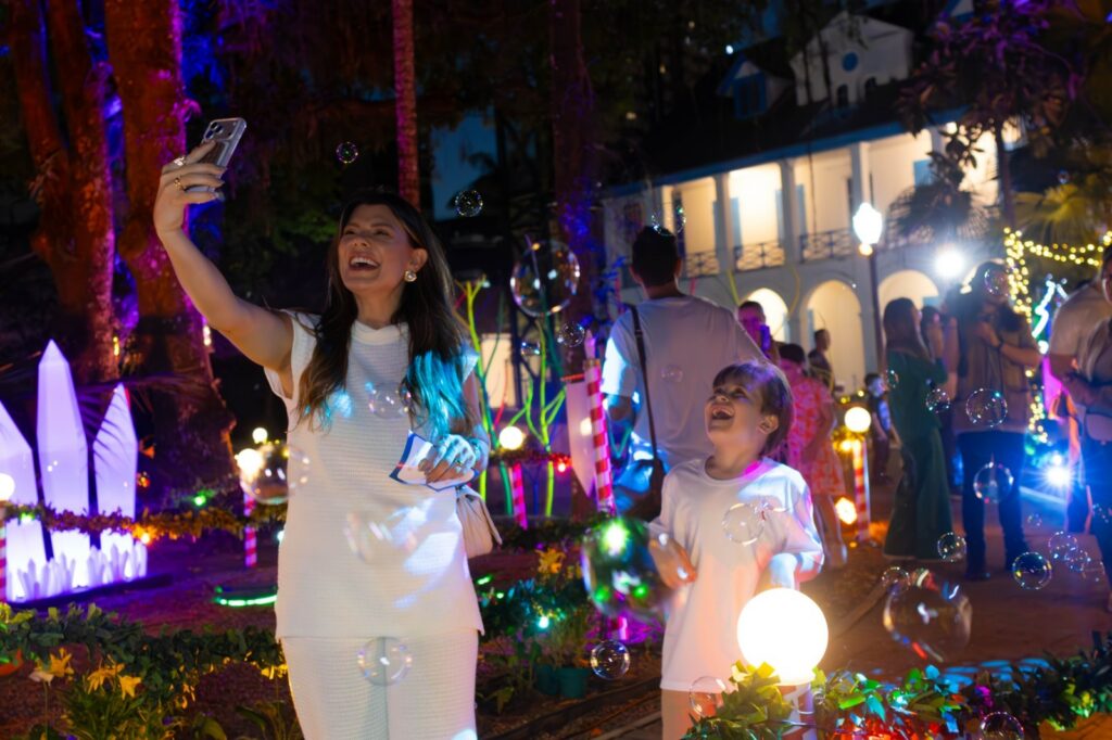 Mulher e menino vestidos de branco sorriem para foto no celular dela enquanto são envoltos por luzes e bolhas de sabão