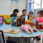 Crianças brincam ao redor de mesa com livros, boneca e outros brinquedos