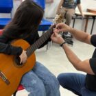 Menina está sentada com violão no colo enquanto professor, ao lado dela, faz ajustes na posição da mão da criança