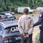 Homem com uniforme da Guarda Municipal segura o braço de outro homem algemado. Os dois estão de costas para a câmera, com uma viatura da Guarda Municipal no fundo da imagem