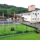 Imagem mostra prédio bege de dois andares com quadra esportiva coberta ao lado e espaços com gramado