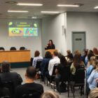 Mulher está em púlpito e fala para plateia que está sentada em cadeiras numa sala ampla