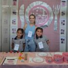 Professora e duas alunas uniformizadas posam para foto no estande do projeto menu da matemática, com decoração rosa