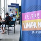 Banner com a frase Feirão Limpa Nome está me primeiro plano. No fundo, pessoas sentadas recebem atendimento
