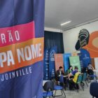 Banner escrito Feirão Limpa Nome Joinville em primeiro plano, com pessoas sentadas em atendimento ao fundo