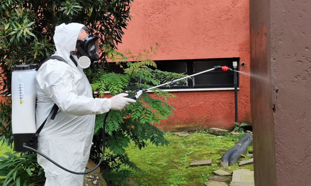 Homem usa roupa de proteção branca enquanto aponta vara de pulverizador para parede