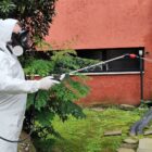 Homem usa roupa de proteção branca enquanto aponta vara de pulverizador para parede