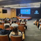 Em auditório, homem fala em pé, no palco, enquanto outras pessoas sentadas assistem à apresentação