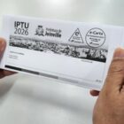 Mãos de um homem seguram folha branca dobrada com o brasão da Prefeitura de Joinville e o selo IPTU 2026