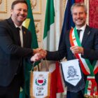 Dois homens de terno apertam as mãos enquanto seguram flâmulas com os símbolos de duas cidades