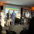 Homens e mulheres estão em pé, em um palco, com telão ao fundo