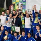 Adolescentes uniformizados com roupa azul sorriem e seguram medalhas, enquanto homens em pé sorriem e levantam troféu