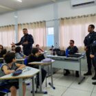 Guardas municipais uniformizados estão em pé, em sala de aula, enquanto alunos estão sentados olhando para os agentes