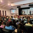 Homem com microfone está em pé, em um palco, enquanto dezenas de pessoas sentadas assistem à apresentação