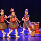Cinco meninas com o mesmo figurino com vestido e laço na cabeça dançam em palco