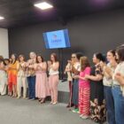 Dezenove mulheres em pé, lado a lado, olham para plateia, batendo palmas