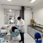 Homem com jaleco branco está em pé manuseando instrumento, enquanto mulher está deitada na cadeira em consultório odontológico