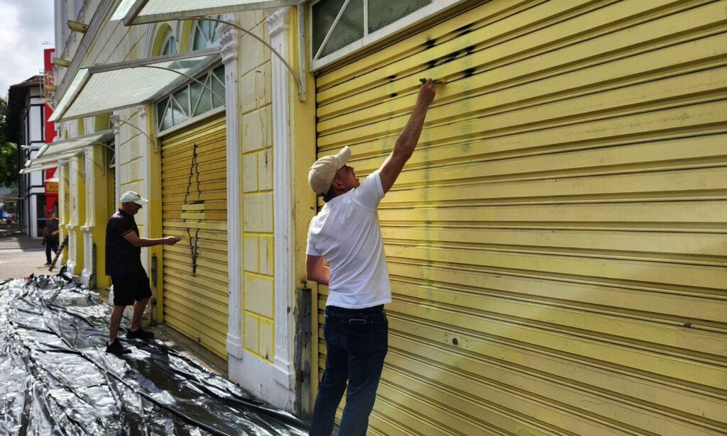 Dois homens estão com pincéis das mãos e passam o acessório com tinta amarela em uma porta