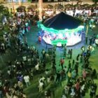 Imagem aérea mostra carrossel iluminado em uma praça com dezenas de pessoas caminhando ao redor do brinquedo