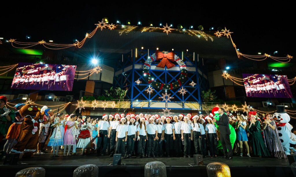 Dezenas de pessoas sorriem em pé no palco, em frente à prédio da Prefeitura iluminado