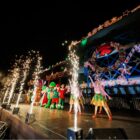 Pessoas com figurino colorido em cima de palco iluminado com efeitos em frente à prédio decorado