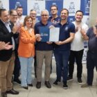 Homens e mulheres estão em pé, sorrindo e posando para a foto. No centro, três pessoas mostram um documento