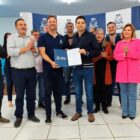 Homens e mulheres estão em pé e sorriem para a câmera. Dois homens seguram um documento