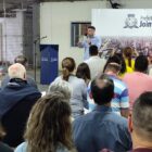 Homem com microfone está em pé em frente à painel com a frase Prefeitura de Joinville enquanto pessoas sentadas acompanham a fala