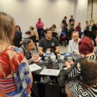 Grupos de pessoas sentados em mesas são observados por outras pessoas em pé