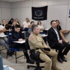 Homem em pé fala para diversas pessoas sentadas em sala