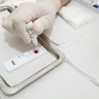 Imagem mostra mão com luva segurando frasco acima de dispositivo com gota de sangue
