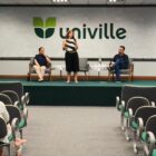 Mulher fala para plateia em palco, segurando microfone. Um homem e uma mulher também estão no palco, sentados em poltronas