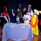 Adolescentes vestidas com diferentes figurinos estão em pé, próximos a uma mesa