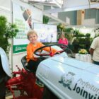 Menino está em cima de trator e segura volante do veículo em estande de Festa das Flores