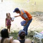 Agente da Defesa Civil entrega material educativo para menina na margem de um rio