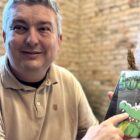 Homem de camisa bege segura livro e aponta o dedo para a capa onde está estampado um desenho de jacaré