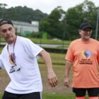 Em um espaço ao ar livre, homem de camisa branca segura bola pequena e se prepara para jogá-la. Ao fundo, outro homem de camisa laranja sorri