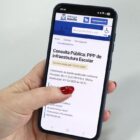Mão feminina segura celular aberto na página da consulta pública online sobre o projeto de Parceria Público-Privada (PPP) de infraestrutura para 27 novas unidades escolares