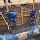 Dois trabalhadores usam pás para movimentar terra dentro de um grande buraco onde está a adutora, um grande tubo azul