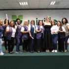 Catorze mulheres estão em um palco, em pé, lado a lado e sorriem para a câmera enquanto seguram um papel