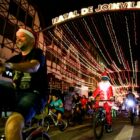 Ciclistas pedalam embaixo de túnel de luzes com escrito "Natal de Joinville". Um deles está vestido de Papai Noel e outro usa gorro